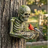 A weathered skull and skeleton sculpture made of resin attached to a tree trunk, with a small bird perched nearby.