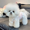 A fluffy white dog wearing a floral print vest sitting on a dark gray cushion.