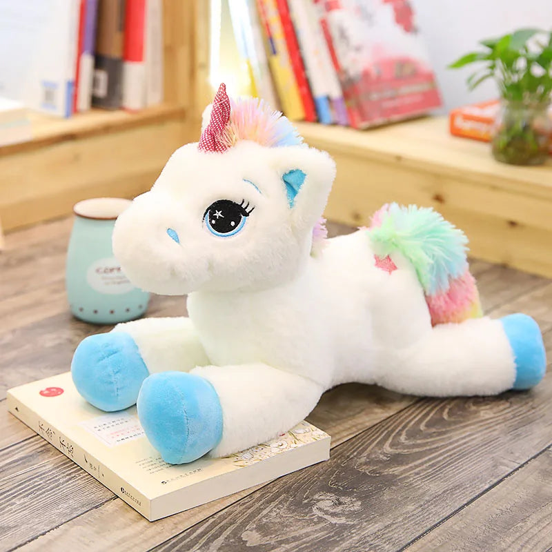 A white unicorn plush toy with pink and blue accents is displayed on a wooden surface next to books.