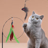 A gray cat is sitting next to an interactive teaser wand with a feather bird and bell hanging from a line.