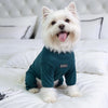 A white dog wearing a dark teal cozy pajama top with a high collar, lying on white bedding.