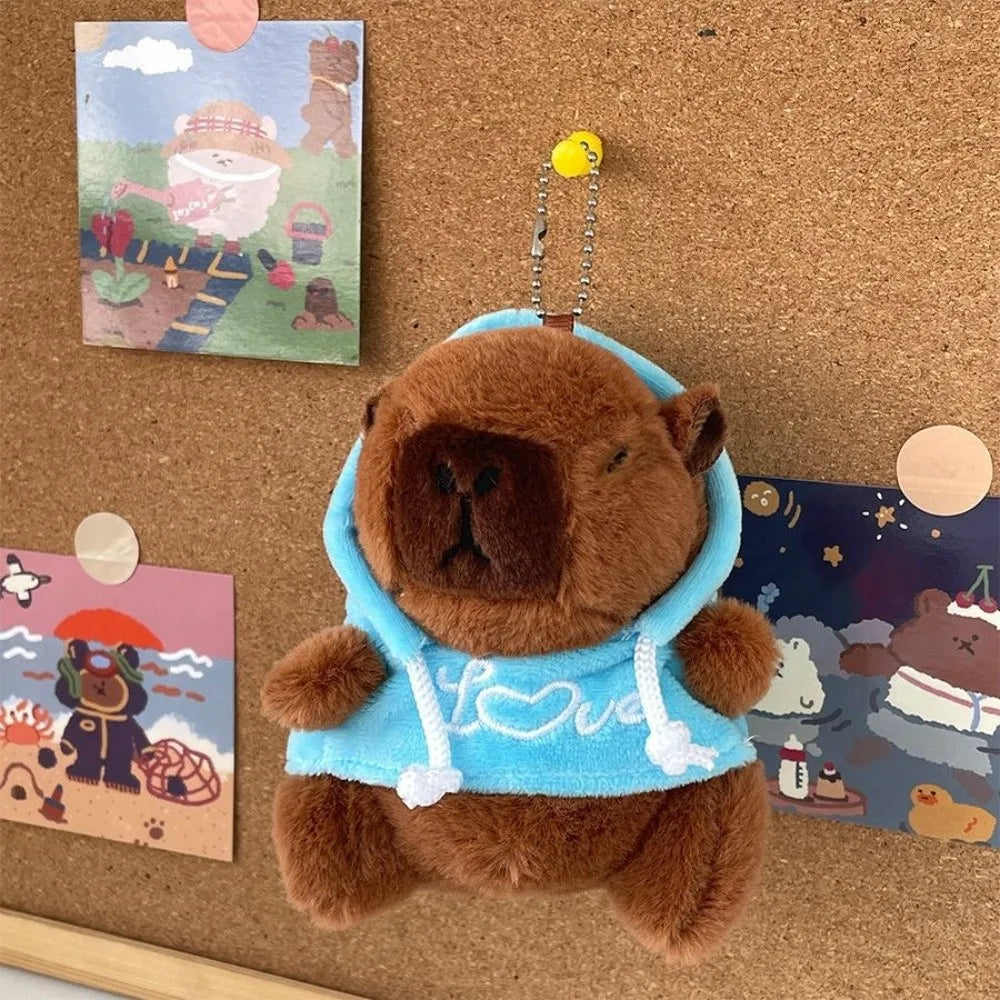 A brown capybara plush hoodie doll with light blue fabric and white trim is displayed on a cork board.