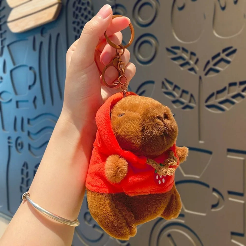 A hand holds a brown capybara plush doll wearing an orange hoodie with small wings against a patterned background.