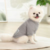 A fluffy white dog wearing a gray knitted winter sweater, lying down on a light surface.