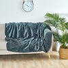 A dark teal microfiber blanket draped over a light-colored sofa with a large green plant in the background.