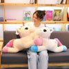A young woman is sitting on a couch holding two white unicorn plush toys with pink and blue accents.