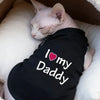 A white cat is resting inside a fluffy brown basket, wearing a black shirt that says I love my Daddy.