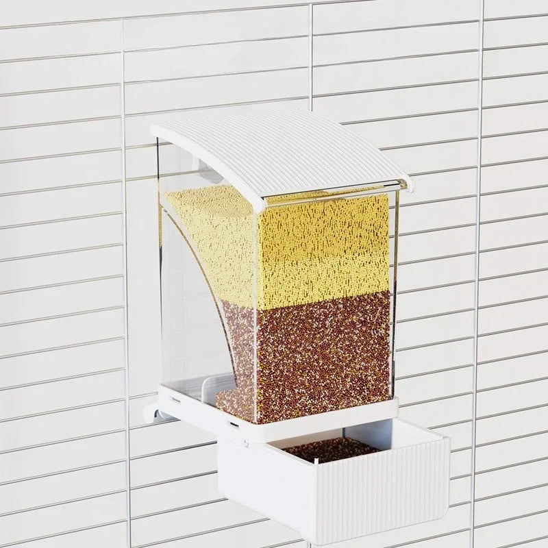 A clear automatic parrot feeder with a white lid, showing layers of seeds and husks inside.