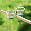 A clear plastic cup feeder with a mounting bracket is attached to a wooden stick, designed for bird feeding.