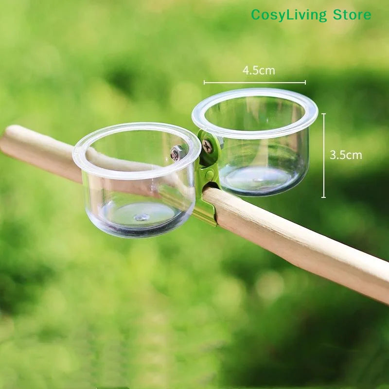 A clear plastic cup feeder with a mounting bracket is attached to a wooden stick, designed for bird feeding.