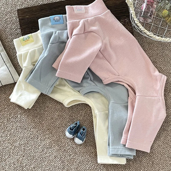 A collection of soft stretch dog pajamas in various colors including pink, gray, and cream, laid out on a brown carpet.