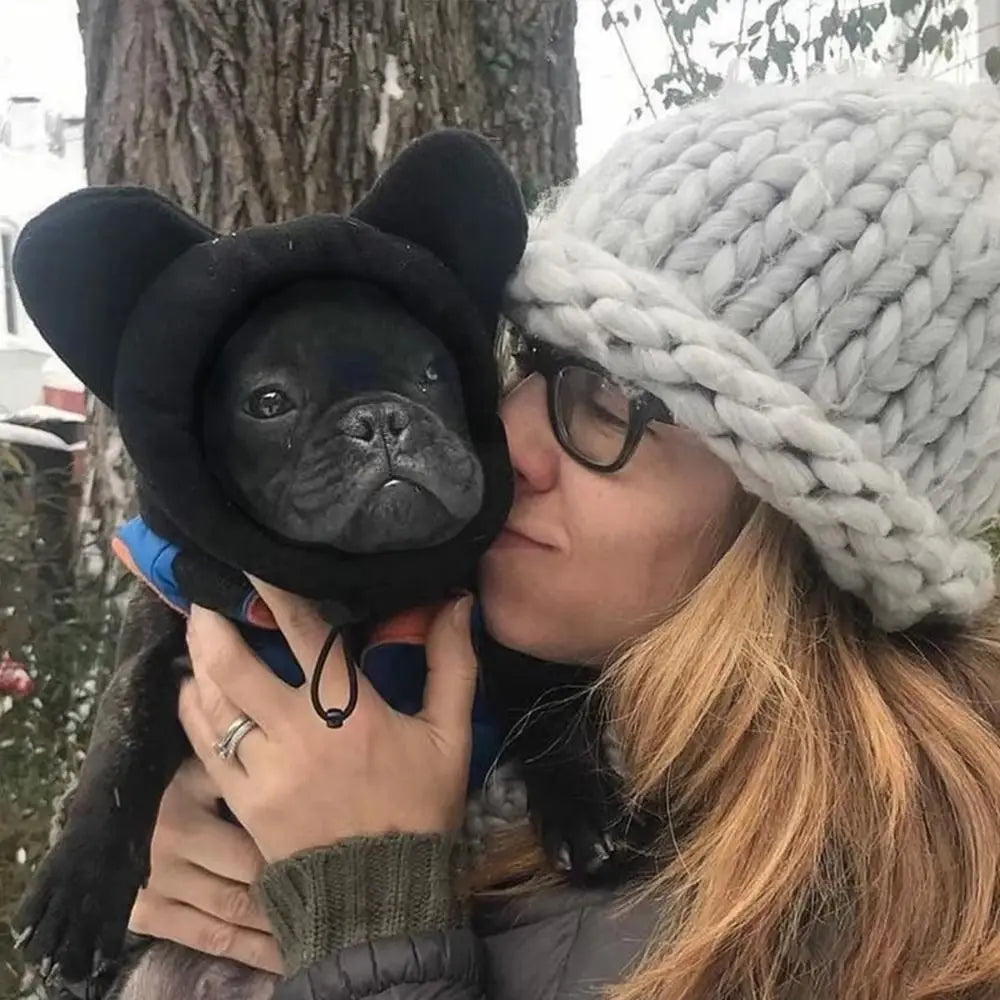 A woman is kissing a black dog wearing a black ears hat while holding a light gray knitted winter hat.