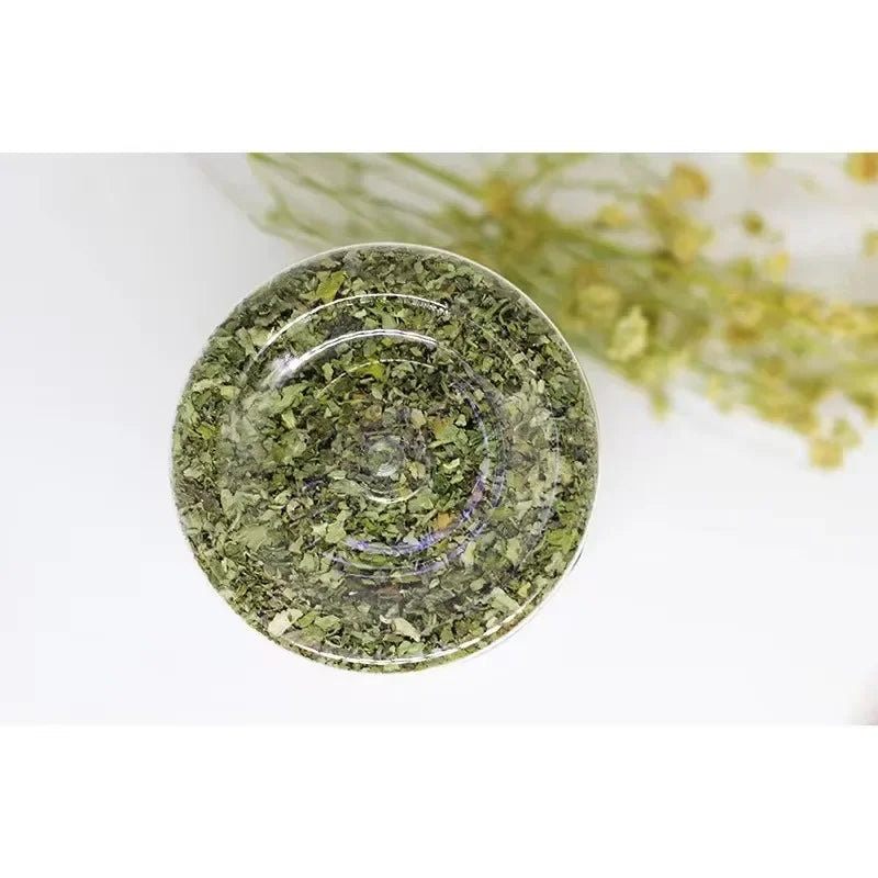 A round, translucent green glass dish filled with dried natural catnip and mint grass for feline relaxation.