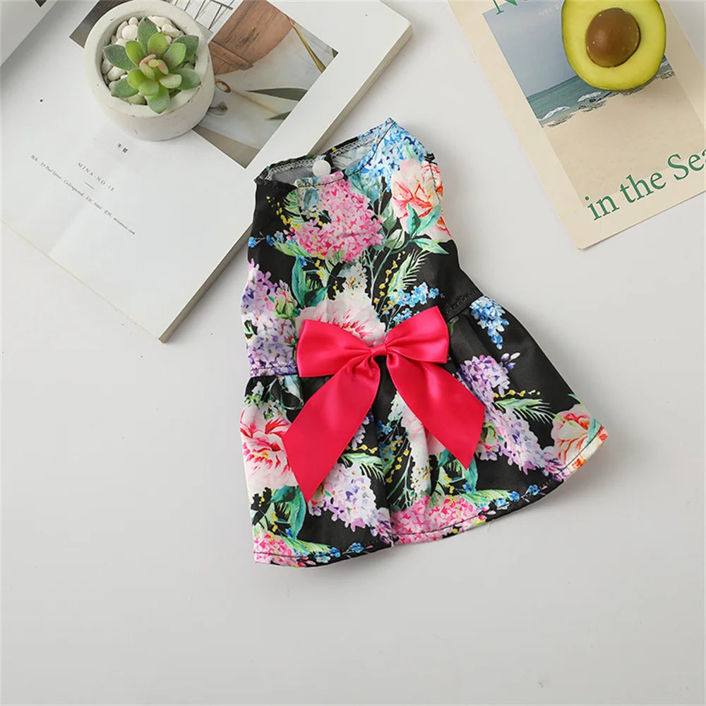 A floral print dog dress with pink and green flowers and a red bow displayed on a white surface.
