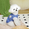 A white poodle wearing a blue striped cooling vest is sitting on a light surface.