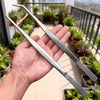 A hand holds a pair of precision stainless steel tweezers, featuring both straight and curved tips.