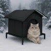 A fluffy gray cat is sitting in a black heated outdoor shelter during a snowy day.