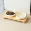 A wooden stand holds two white ceramic bowls, one filled with dark kibble for cats and dogs.