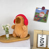 A fluffy brown capybara plush doll wearing a red hood sits next to a small glass of flowers and a framed artwork.