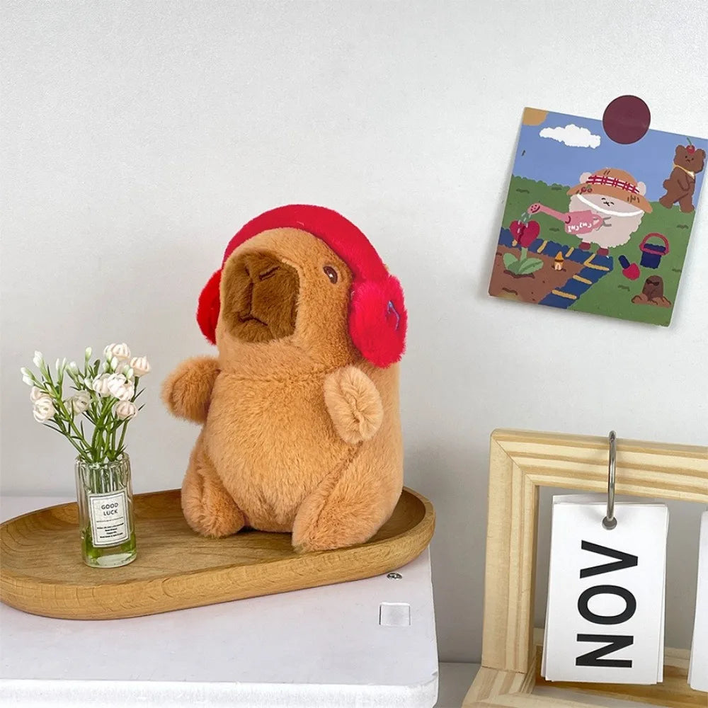 A fluffy brown capybara plush doll wearing a red hood sits next to a small glass of flowers and a framed artwork.