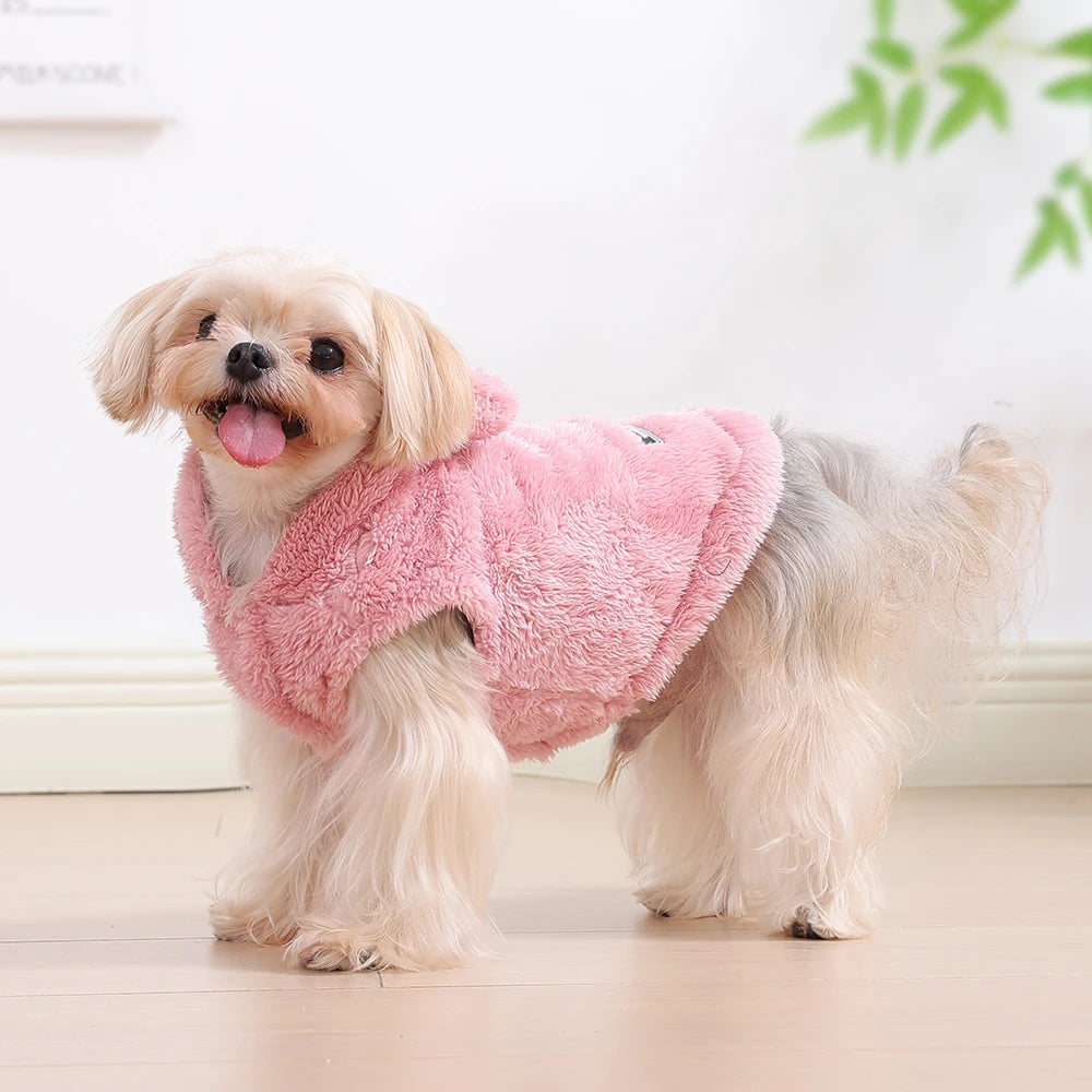 A light tan dog wearing a pink fuzzy fleece hoodie standing on a wooden floor.
