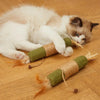 A white kitten is lying on a wooden surface next to several natural wooden teething sticks for cats.