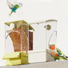 A clear automatic parrot feeder with a yellow base and a separate husk compartment, featuring a small green bird flying above