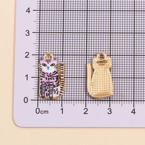 Two gold-toned cat pendant charms are displayed on a grid ruler, showing a purple and black striped cat design.