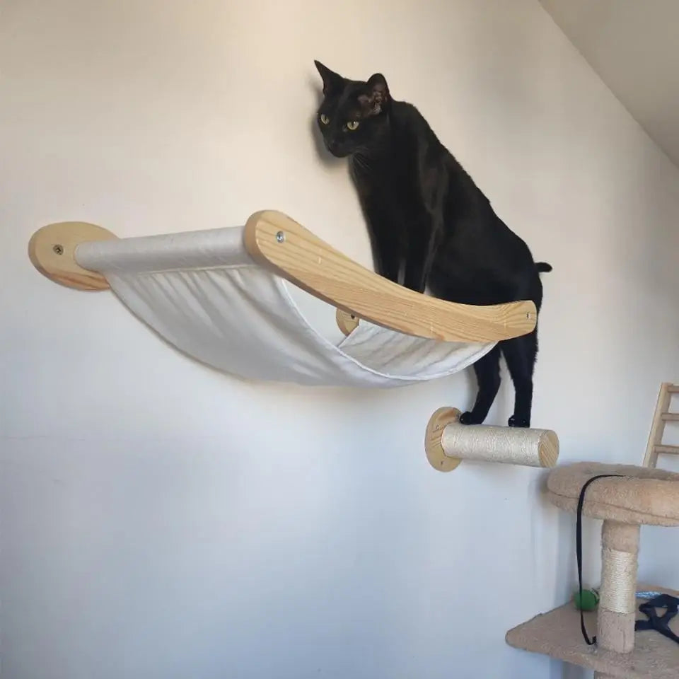 A black cat is perched on a wooden modular cat wall shelf featuring a bridge and hammock.