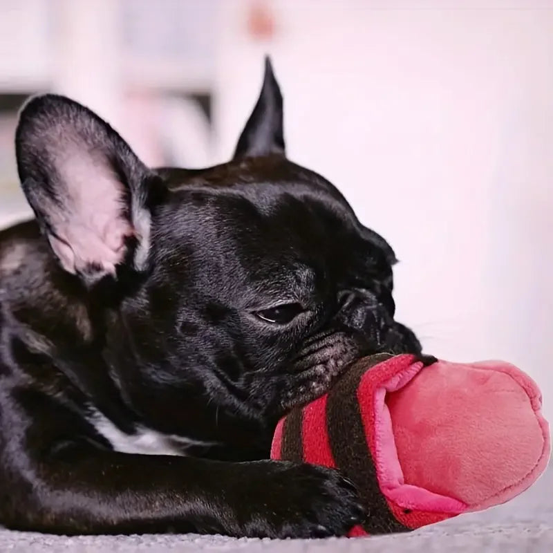 A black French Bulldog puppy is lying down and chewing a pink plush dog chew toy.
