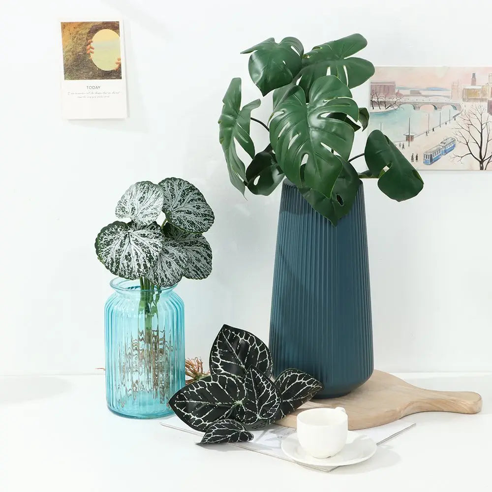 Eine hohe, dunkeltürkise Vase hält eine große grüne Monstera-Blattpflanze auf einer weißen Oberfläche.