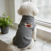 A white dog wearing a gray sleeveless vest with a red and black cartoon embroidery patch.