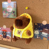 A brown capybara plush doll wearing a yellow hoodie with white trim, set against a cork board.