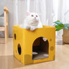 A fluffy white cat is sitting inside a yellow cardboard scratching house with circular cutouts.