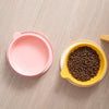 A pink silicone pet feeding plate and a yellow bowl filled with dark kibble on a light wooden surface.