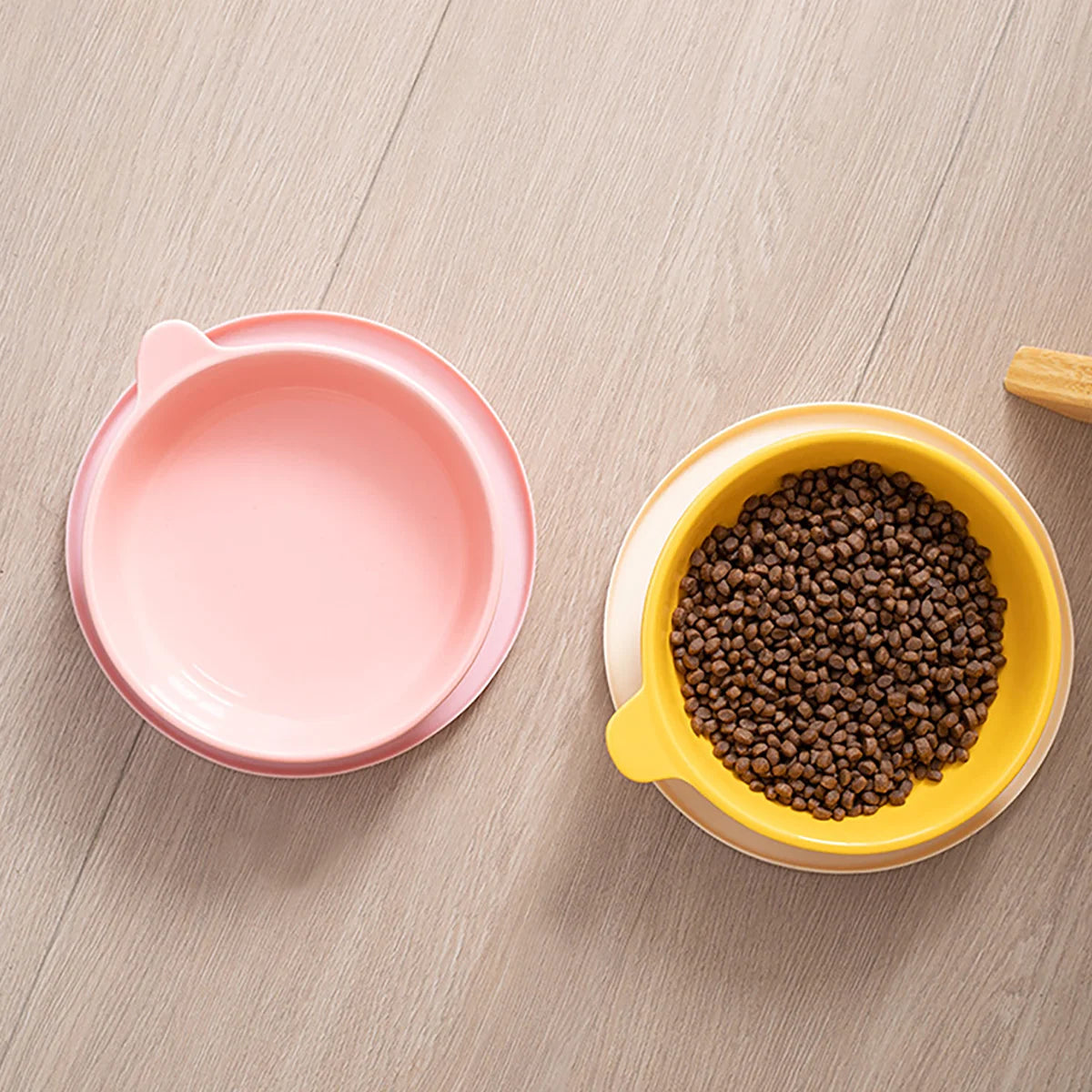 A pink silicone pet feeding plate and a yellow bowl filled with dark kibble on a light wooden surface.