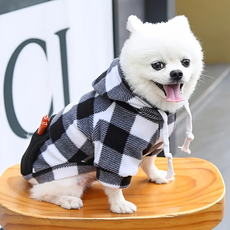 Un perro blanco vistiendo una sudadera con capucha de forro de cuadros negros y blancos, de pie sobre una superficie de mader