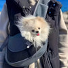 A fluffy white dog is carried in a black pet carrier sling bag by a person.