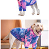 A light-colored dog wearing a tie-dye pink and blue fleece hoodie standing on a floor.