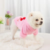 A fluffy white dog wearing a pink hoodie with a red bow, sitting on a white rug.