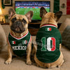 A fawn-colored dog wearing a green and white soccer jersey with the flag of Mexico on the chest, sitting indoors.