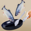 A hand is holding a silver spatula over a black frying pan with an electric floppy fish toy visible.