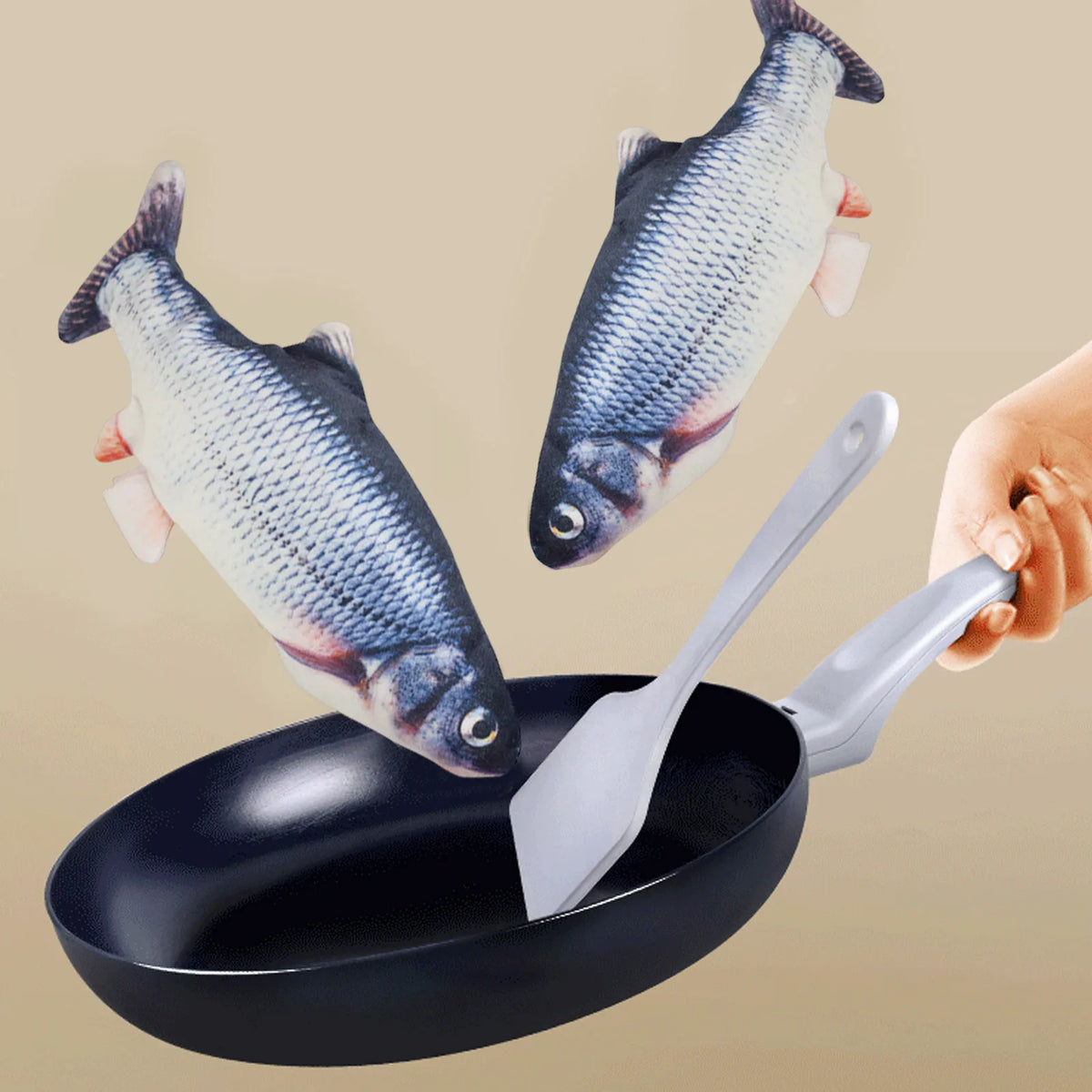 A hand is holding a silver spatula over a black frying pan with an electric floppy fish toy visible.