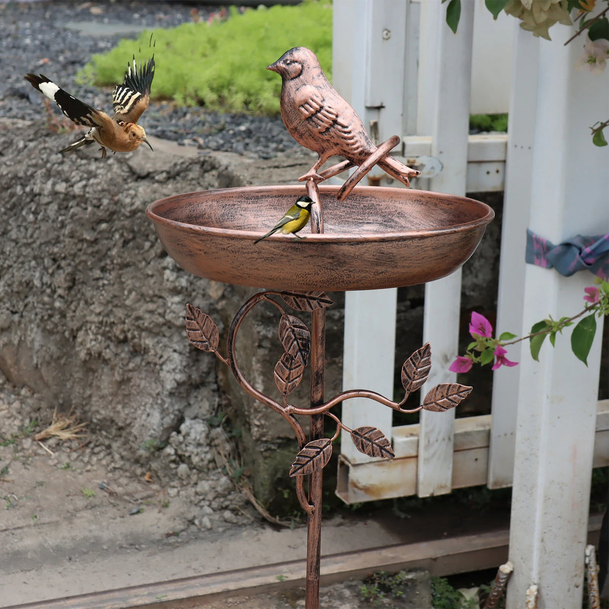 Kupfer-Vogelbad auf einem Ständer mit einem kleinen Vogel darüber, im Freien neben einem weißen Zaun.