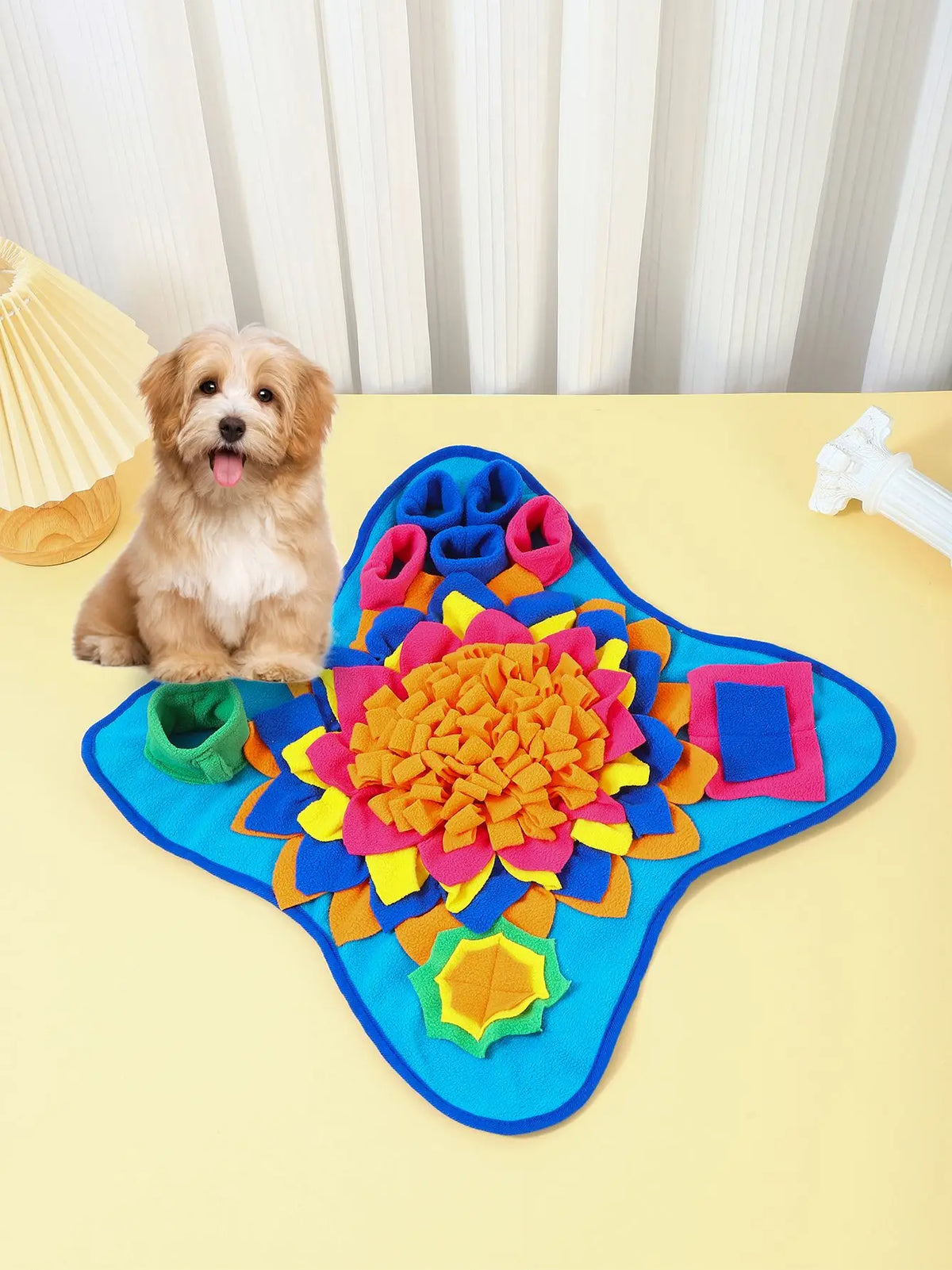 A dog is sitting next to a brightly colored snuffle mat with fabric flowers and puzzle elements on a yellow surface.