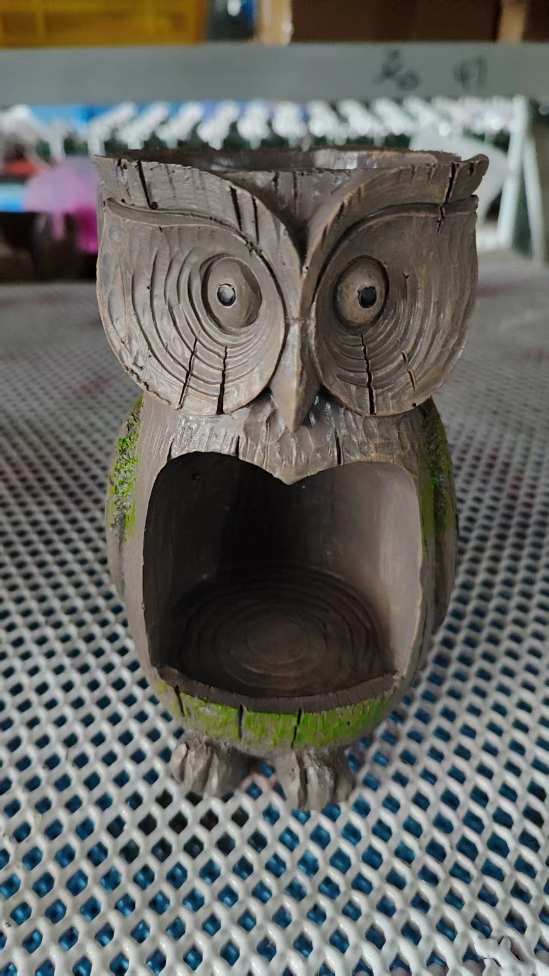 A carved wooden owl bird feeder with a hollow body and green moss accents sits on a blue mesh surface.