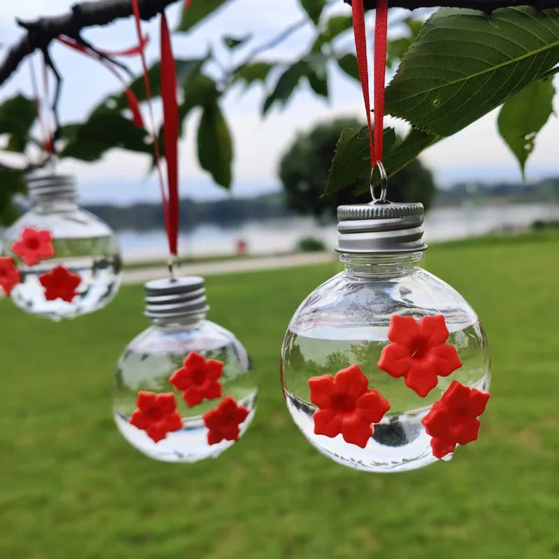 Un distributeur d'eau sphérique en verre clair rempli d'eau et de fleurs rouges, suspendu à une branche.