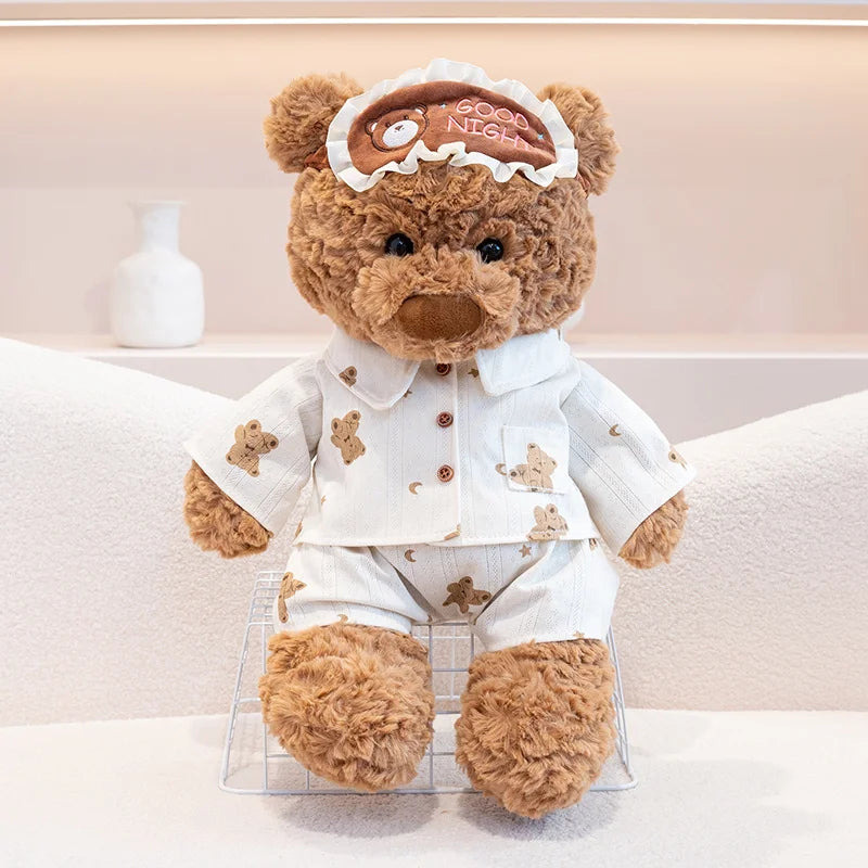 A fluffy brown teddy bear wearing a white collared shirt and pants with an eye mask on its head.
