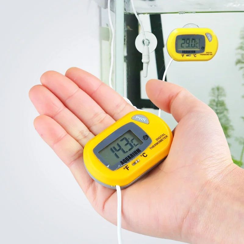 A hand holds a yellow digital thermometer measuring water temperature in an aquarium.