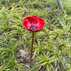 A bright red, flower-shaped stainless steel bird feeder with a black seed dish is planted in green grass.